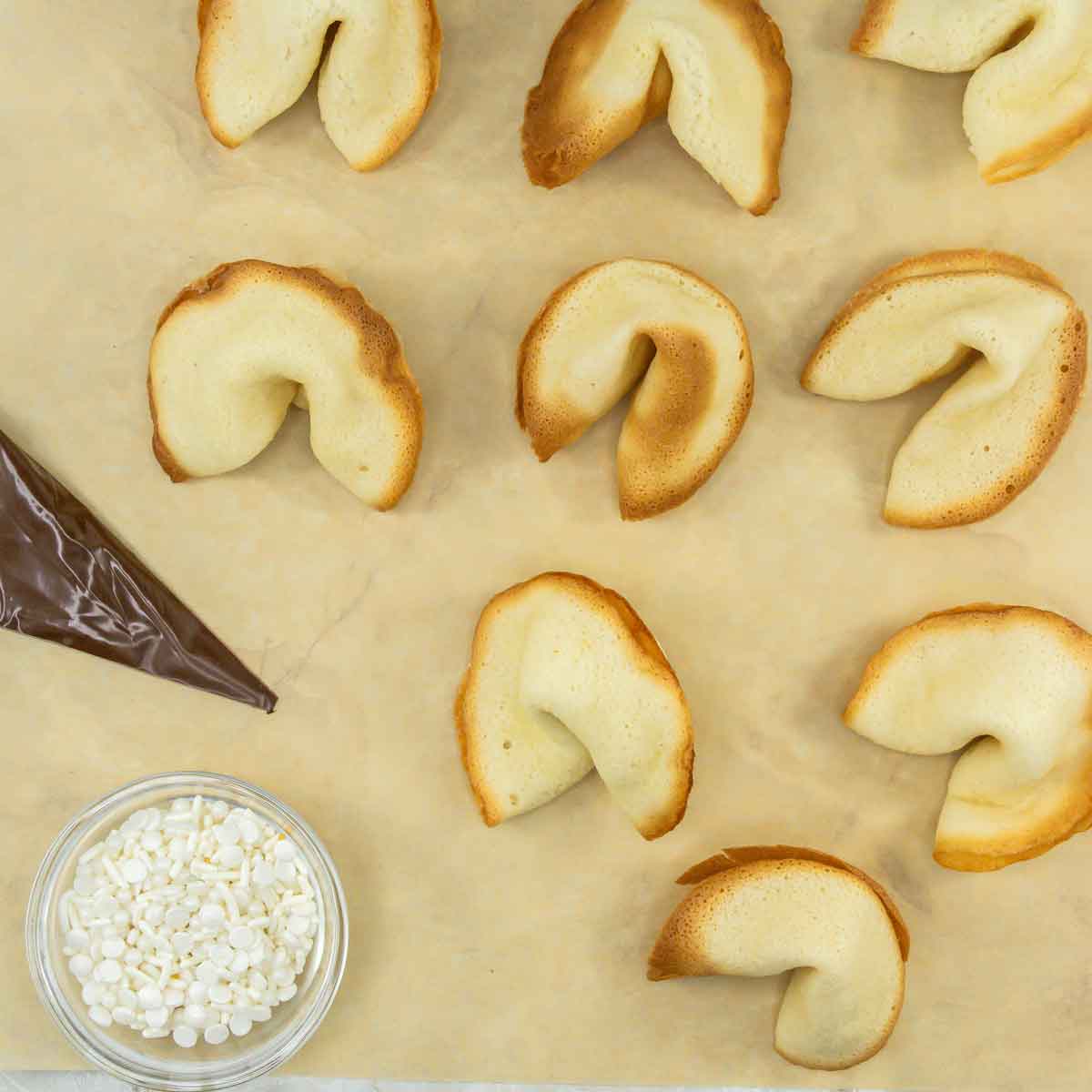A sheet of parchment paper with twelve fortune cookies scattered on it. A piping bag filled with chocolate is on the left, and a small glass bowl with white sprinkles is beside it.