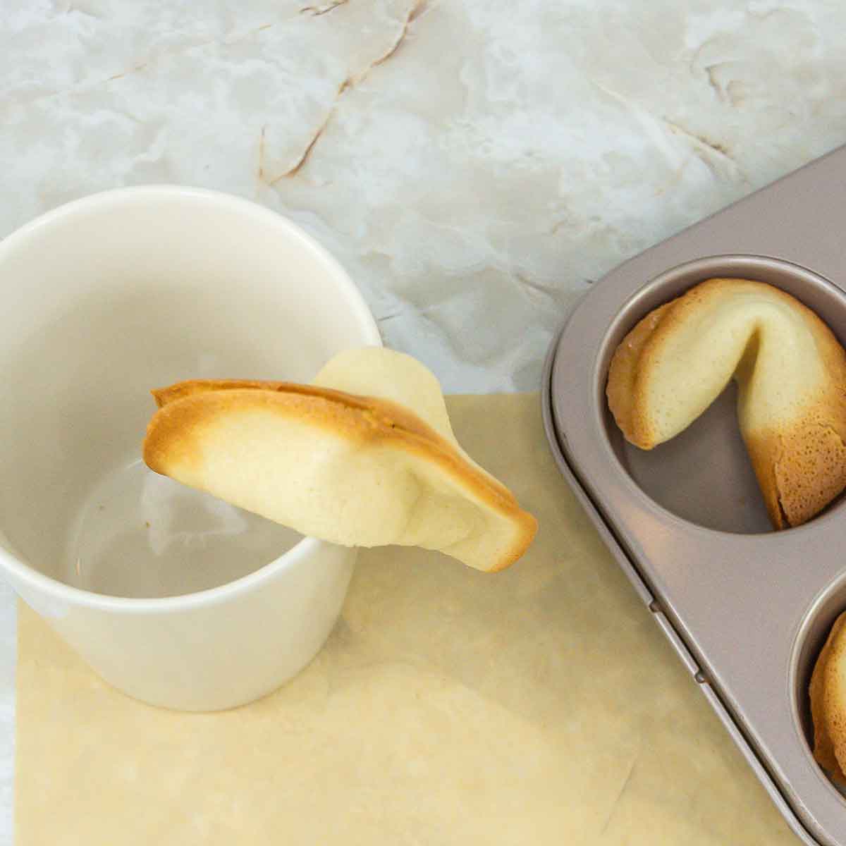A fortune cookie hanging over the rim of a white mug, with an empty muffin tin holding other fortune cookies on a marble countertop.
