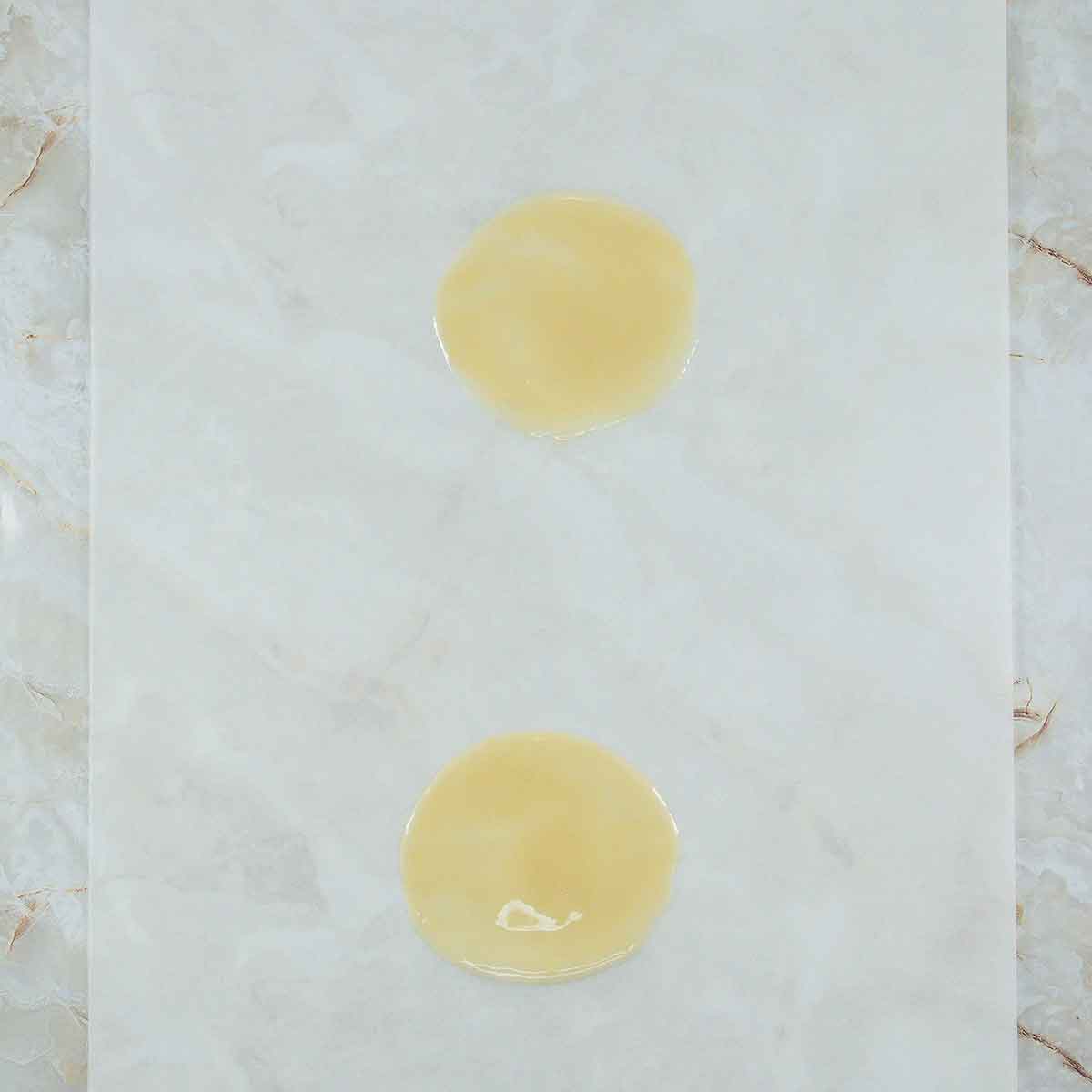 Two round, yellowish liquid stains on a white marble surface, positioned vertically in the center of the image. The texture of the surface is smooth with faint gray veining.