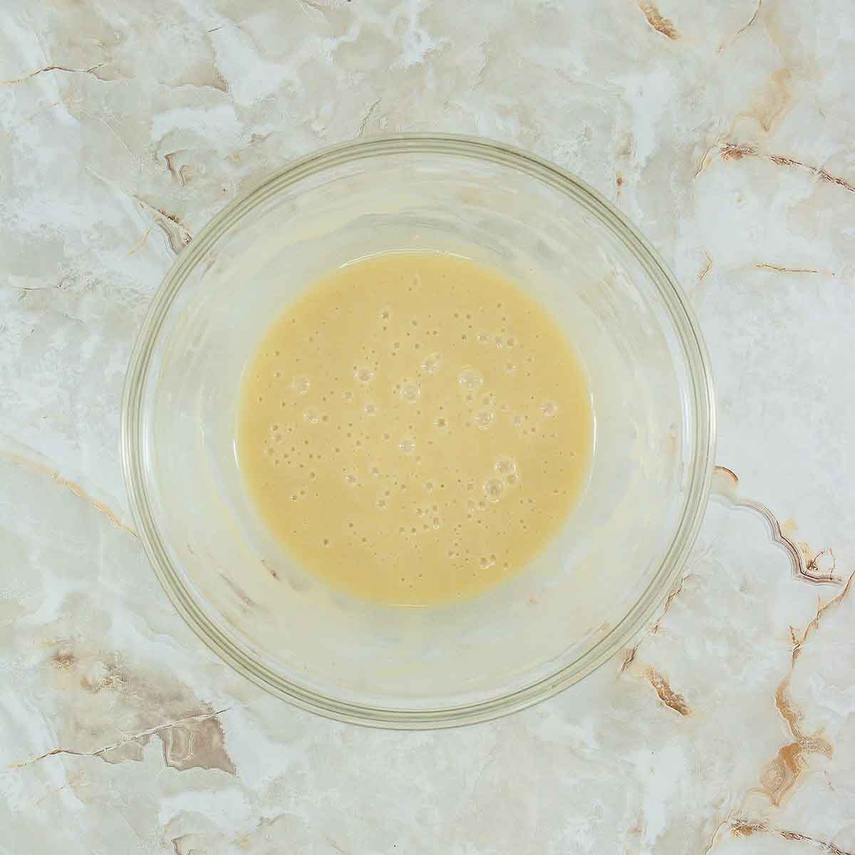 A glass bowl containing a smooth, light beige batter sits on a marbled surface. The batter has a slightly bubbly texture with small air pockets visible.