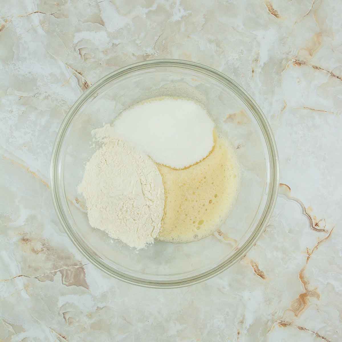 A glass bowl on a marbled surface contains sugar, flour, and a frothy, liquid mixture, possibly eggs being mixed for baking.