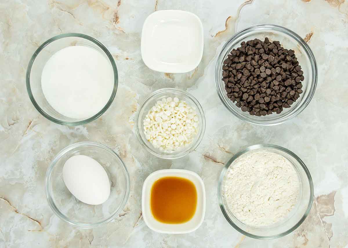 Ingredients on a marble surface: bowls of sugar, chocolate chips, white chocolate chips, flour, vanilla extract, a single egg, and a small dish of salt.