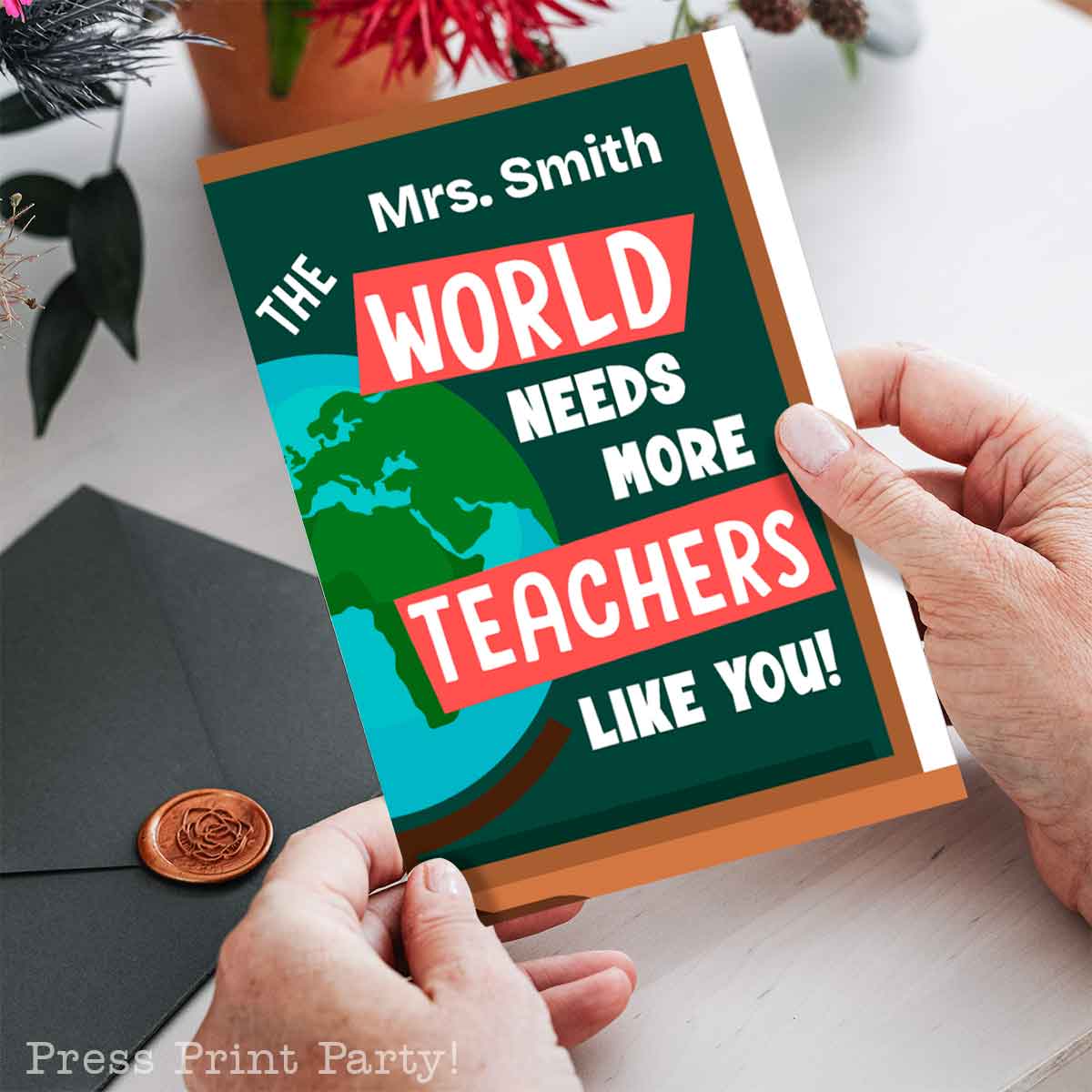 Person holding a card with the text "Mrs. Smith, The World Needs More Teachers Like You!"—a heartfelt birthday wish for teachers. The card boasts a globe design, and nearby an envelope with a wax seal sits beside a plant and festive decoration.