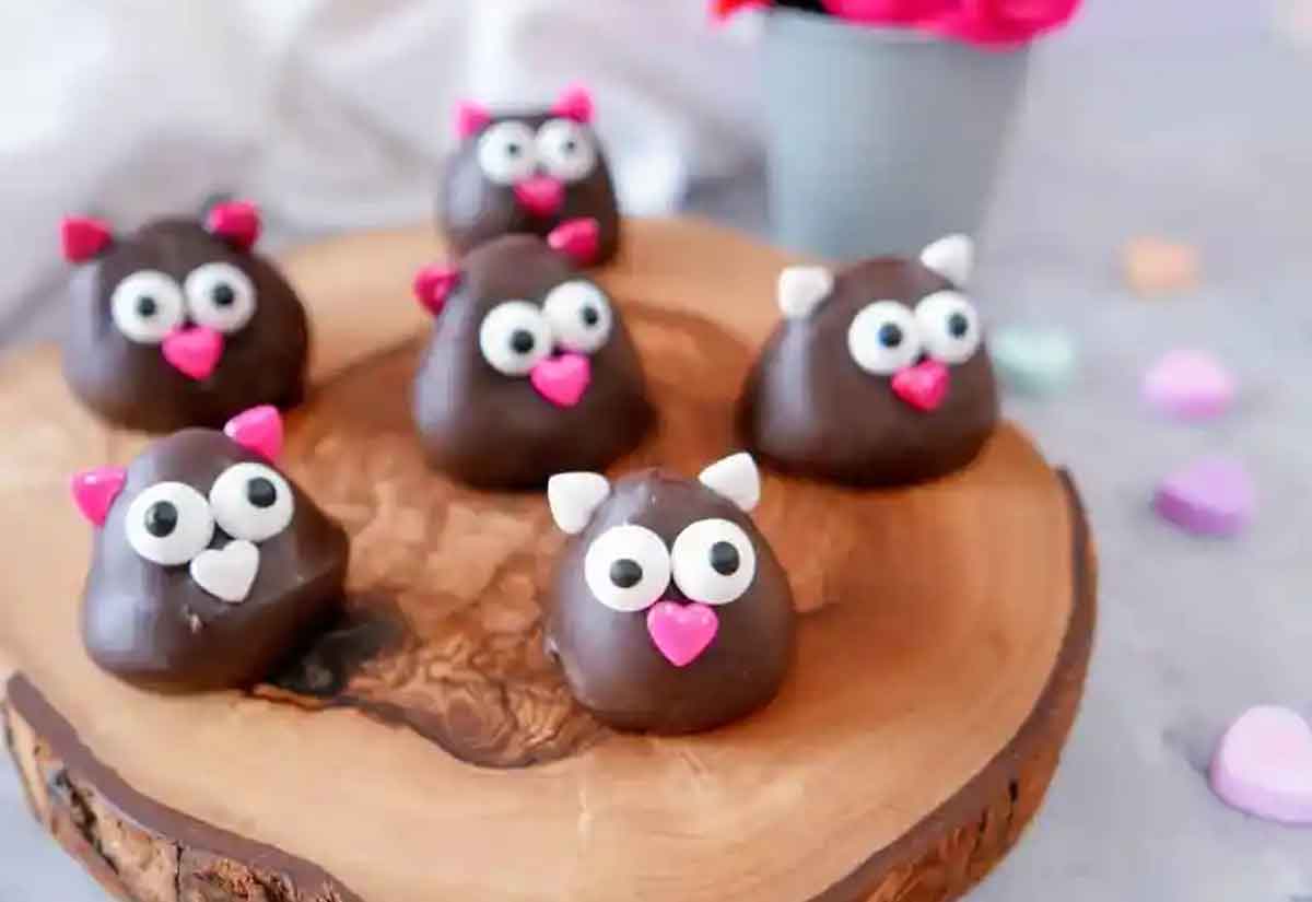 Six charming chocolate confections with large white eyes and pink or white ears sit on a wooden platter, each featuring a pink heart-shaped nose. Perfect healthy Valentine snacks for the classroom, they delight against the backdrop of a blurred potted plant with pink flowers.