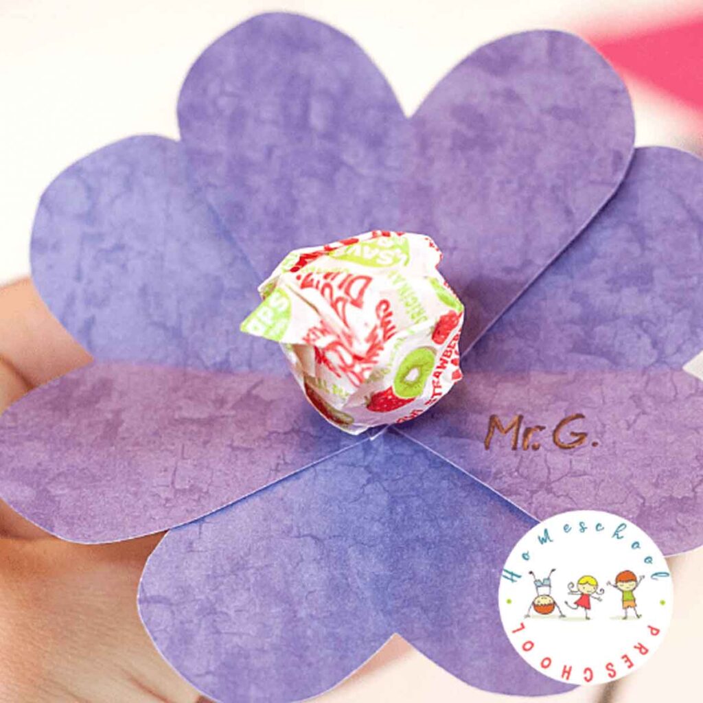 A hand holds a paper flower with purple petals and a candy in the center. The initials "M.G." are written on one petal. A logo with cartoon children and "Preschool Homeschool" text is in the corner.