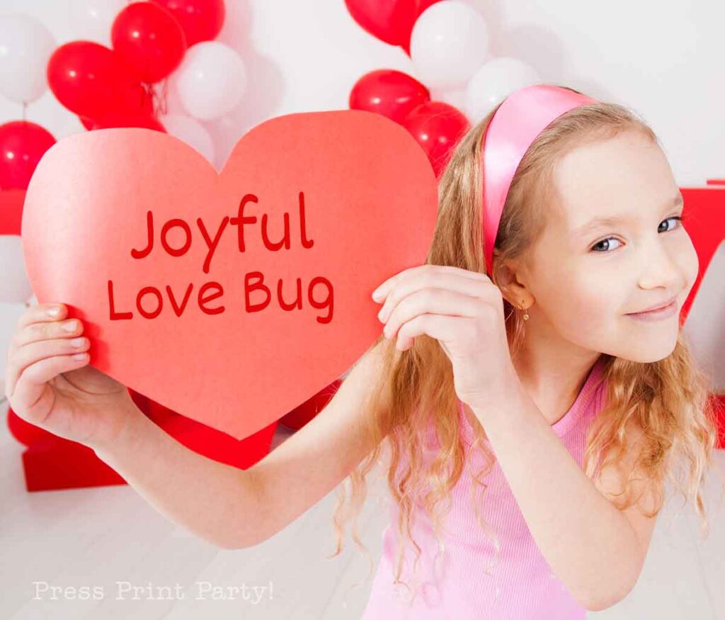 A girl with long blonde hair and a pink headband holds a heart-shaped red sign that reads "Joyful Love Bug," perfect for Valentines Day names. She's wearing a light pink shirt, surrounded by festive red and white balloons.