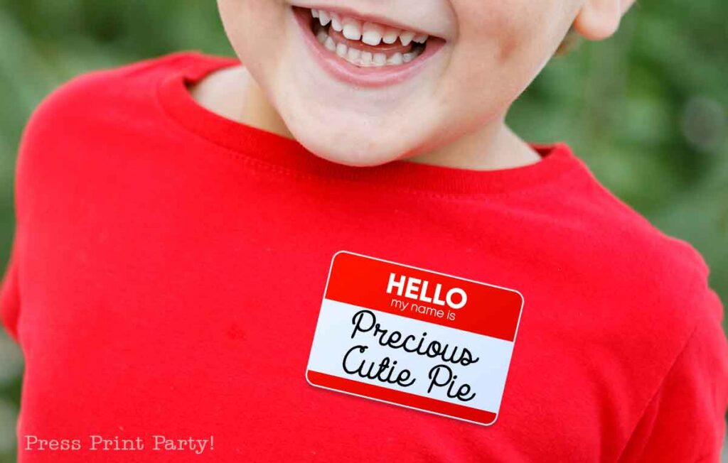 A child wearing a red shirt is smiling. On the shirt, there is a name tag sticker that reads "Hello, my name is Precious Cutie Pie," perfect for Valentine's Day names.