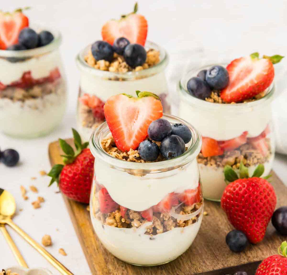 Four glass jars filled with yogurt, granola, and topped with fresh strawberries and blueberries are displayed on a wooden board. Perfect as healthy Valentine snacks for the classroom, additional strawberries and blueberries are scattered around, with two gold spoons nearby.