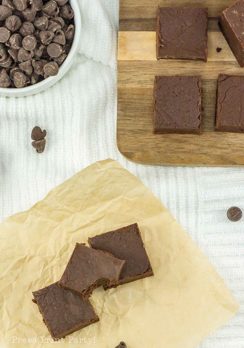 Chocolate fudge squares on a parchment paper and wooden board, surrounded by chocolate chips. A bowl filled with chocolate chips is placed nearby on a white surface.