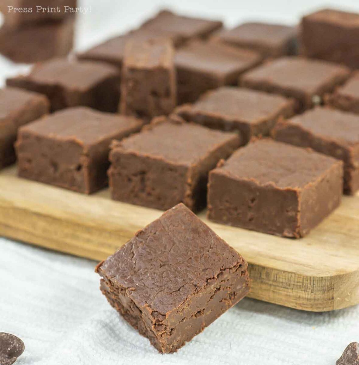 Close-up of several pieces of chocolate fudge arranged on a wooden board. One piece is positioned in front of the board. The fudge has a rich, smooth texture and a deep brown color.