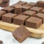 Close-up of several pieces of chocolate fudge arranged on a wooden board. One piece is positioned in front of the board. The fudge has a rich, smooth texture and a deep brown color.