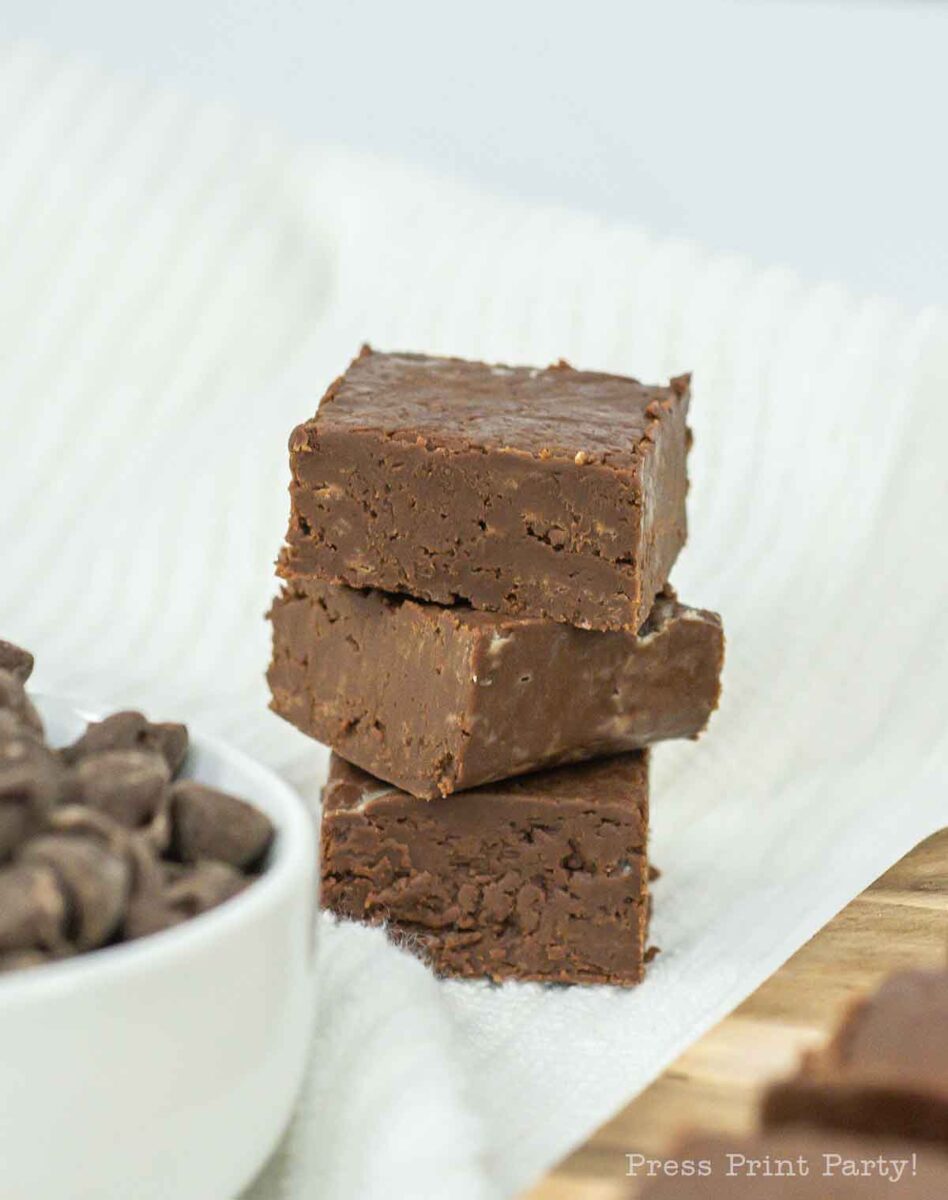 Three squares of chocolate fudge are stacked on a white cloth with a bowl of chocolate chips nearby, set on a wooden board.