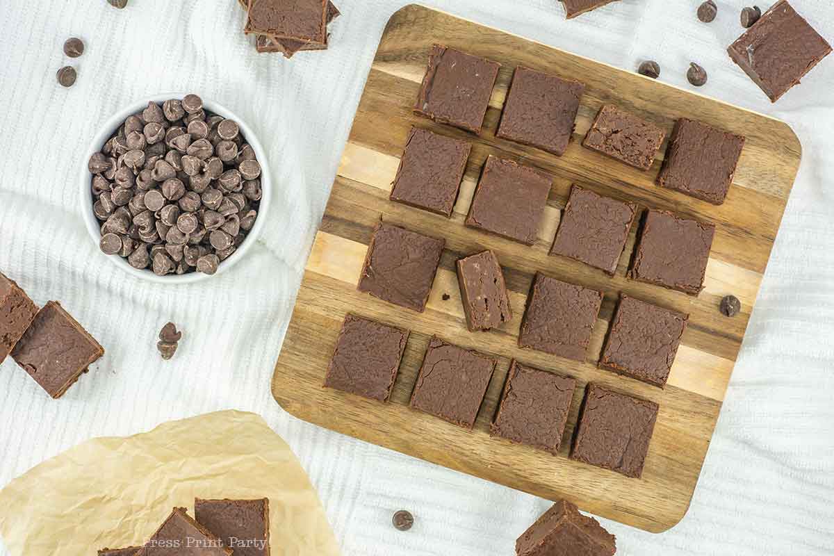 A wooden board with neatly arranged chocolate brownies, two stacked in the center. A bowl of chocolate chips is beside the board, scattered chips around. Some brownies on parchment paper. Set on a white textured background.