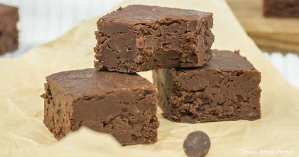Three pieces of rich, dense chocolate fudge stacked on a piece of parchment paper. One piece is on top and two are side by side on the bottom, showcasing their textured surface and delicious appearance. A small chocolate chip is nearby.