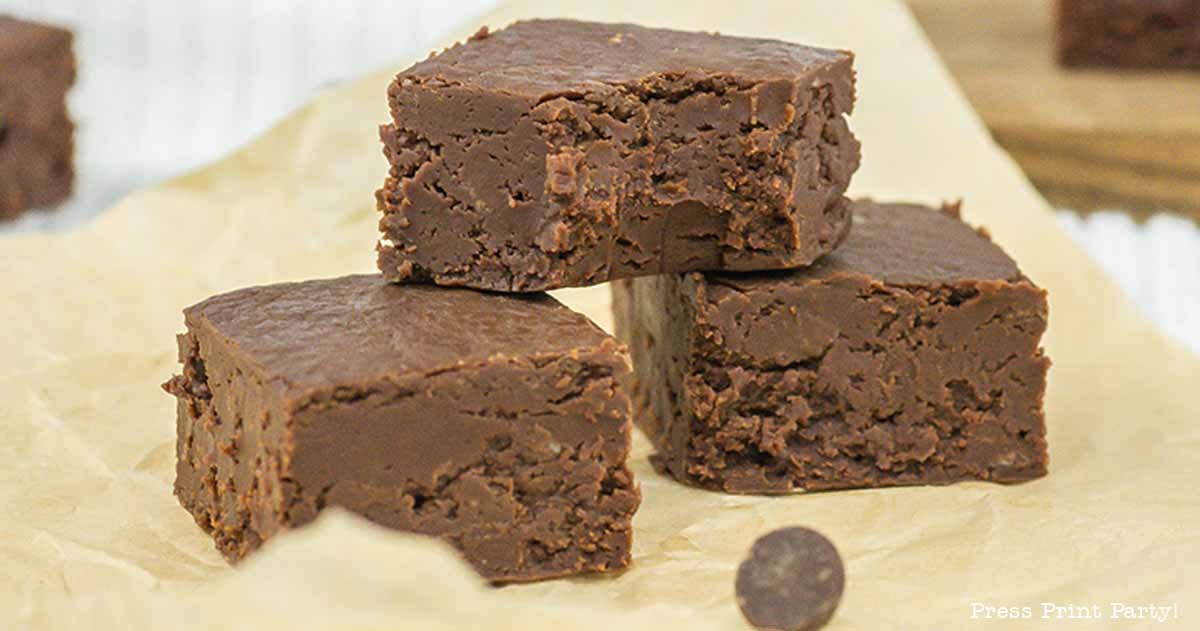 Three pieces of rich, dense chocolate fudge stacked on a piece of parchment paper. One piece is on top and two are side by side on the bottom, showcasing their textured surface and delicious appearance. A small chocolate chip is nearby.