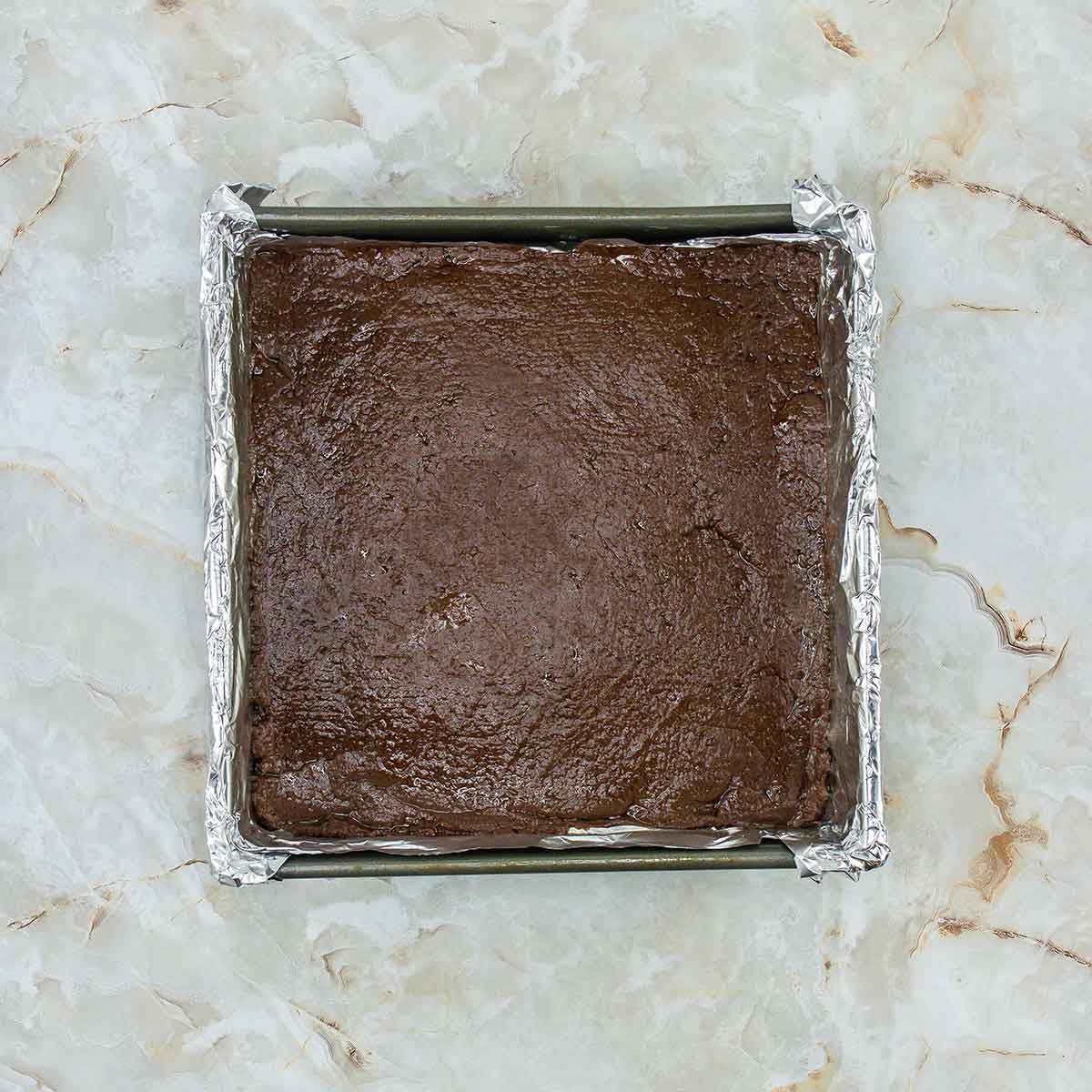 A square baking pan lined with foil, filled with unbaked brownie batter. The pan is placed on a marble countertop.