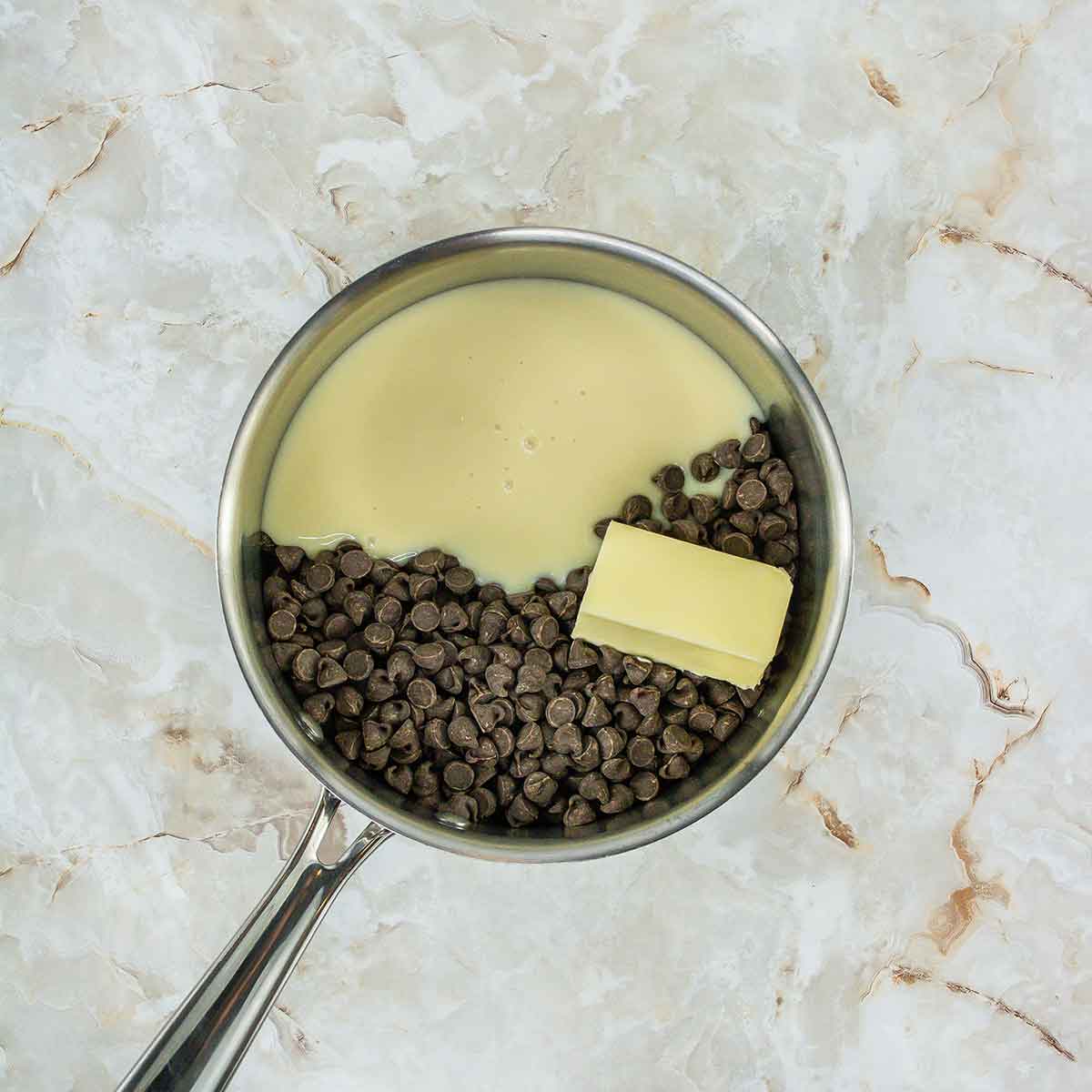 A saucepan on a marbled surface contains chocolate chips, a block of butter, and creamy condensed milk, ready to be melted and mixed together.