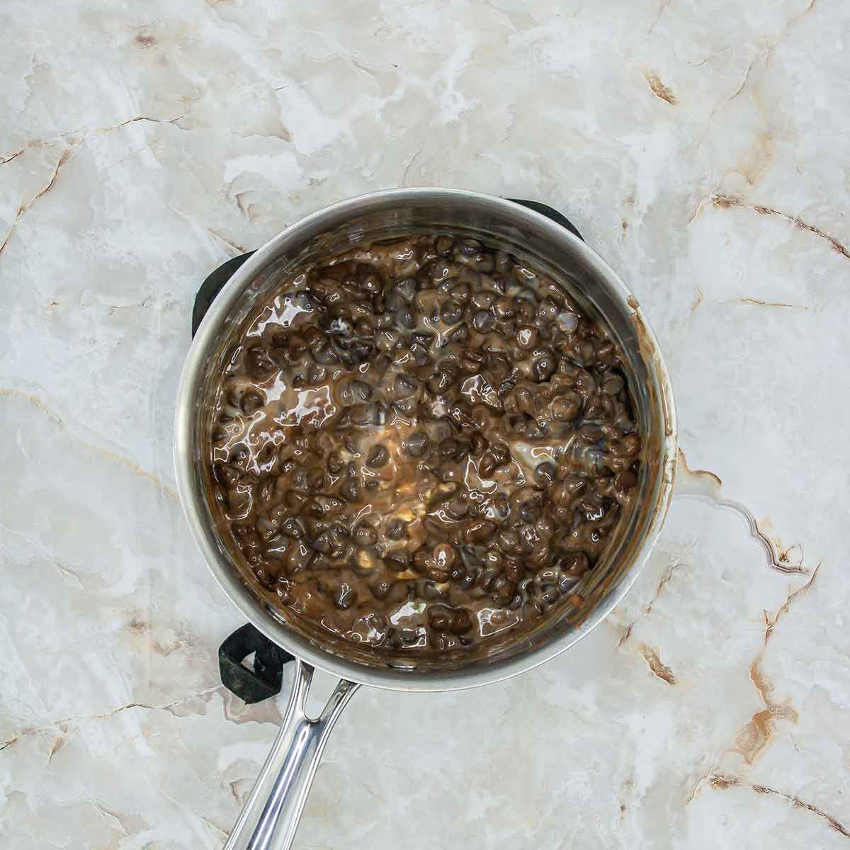 A silver pot filled with cooked black beans on a marble countertop. The beans are submerged in a thick, dark broth.