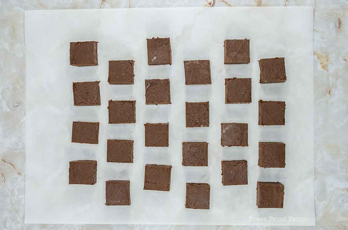 Twenty-five square pieces of chocolate fudge evenly arranged on a sheet of parchment paper. The background is a light-colored marble surface.