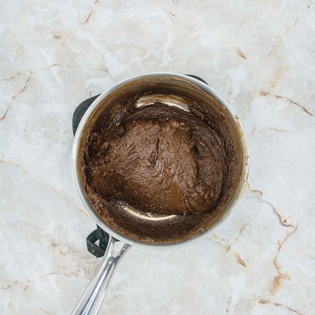 A pot of cooked, thick chocolate ganache sits on a marble countertop. The ganache has a smooth, glossy texture. The pot has a metallic handle and the marble has a light, textured pattern.