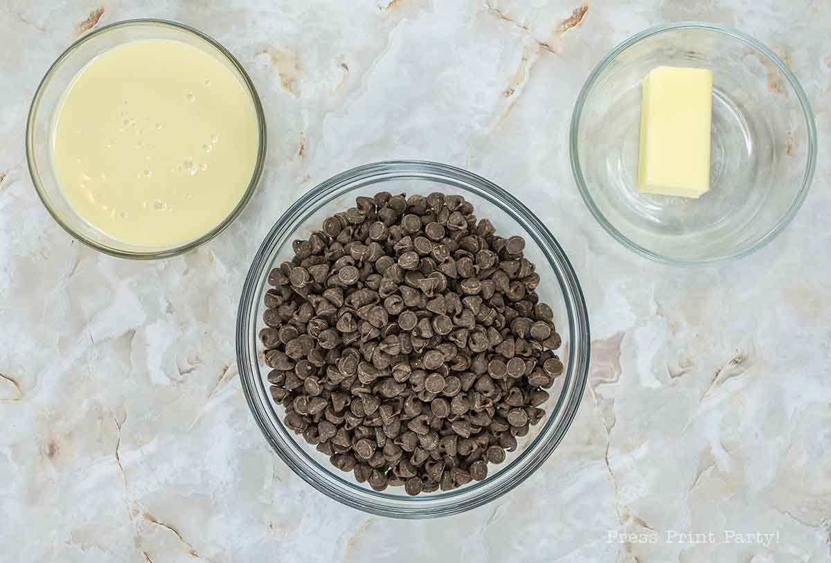 A top-down view of a marble countertop with three glass bowls: one with chocolate chips, another with a stick of butter, and the third containing condensed milk.