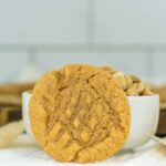 A close-up of a 3 Ingredient Peanut Butter Cookie with a crisscross pattern rests against a white bowl filled with peanuts. The background features a blurred kitchen setting, creating a warm and inviting atmosphere.