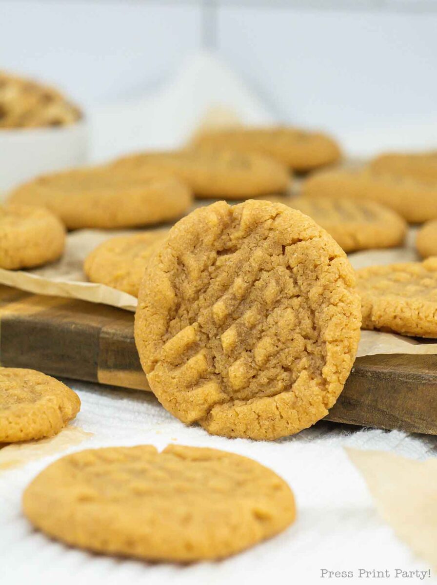 A close-up view of freshly baked 3 ingredient peanut butter cookies, one poised upright on a wooden board amid others lying flat. The cookies boast distinctive fork marks and a soft, crumbly texture. A white bowl of peanuts is visible in the background.