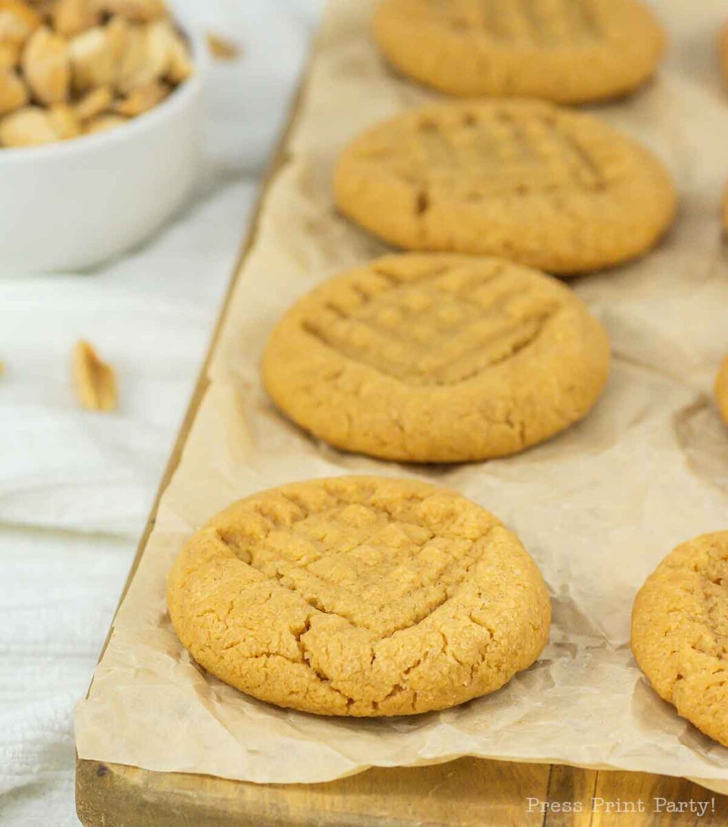 Freshly baked 3 ingredient peanut butter cookies with a classic criss-cross pattern on top are arranged on a wooden board lined with parchment paper. A bowl of peanuts is visible in the background, adding to the cozy homemade vibe.