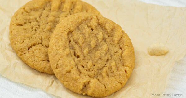 Two delightful three-ingredient peanut butter cookies with fork impressions rest on crumpled parchment paper. A small dollop of peanut butter is nearby, adding a homemade charm.