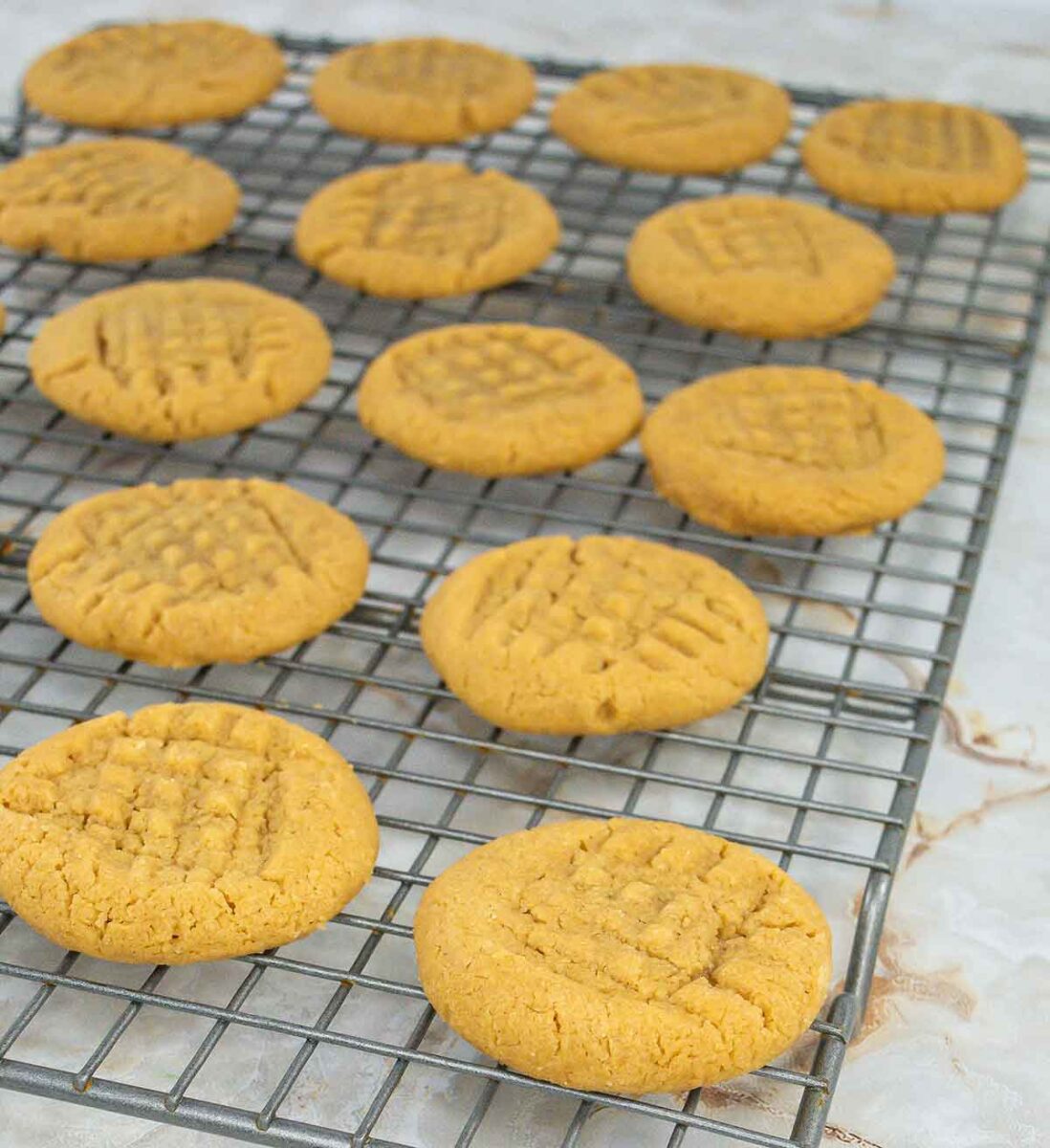 Freshly baked 3-ingredient peanut butter cookies with crisscross fork patterns cool on a metal wire rack placed on a marble surface.