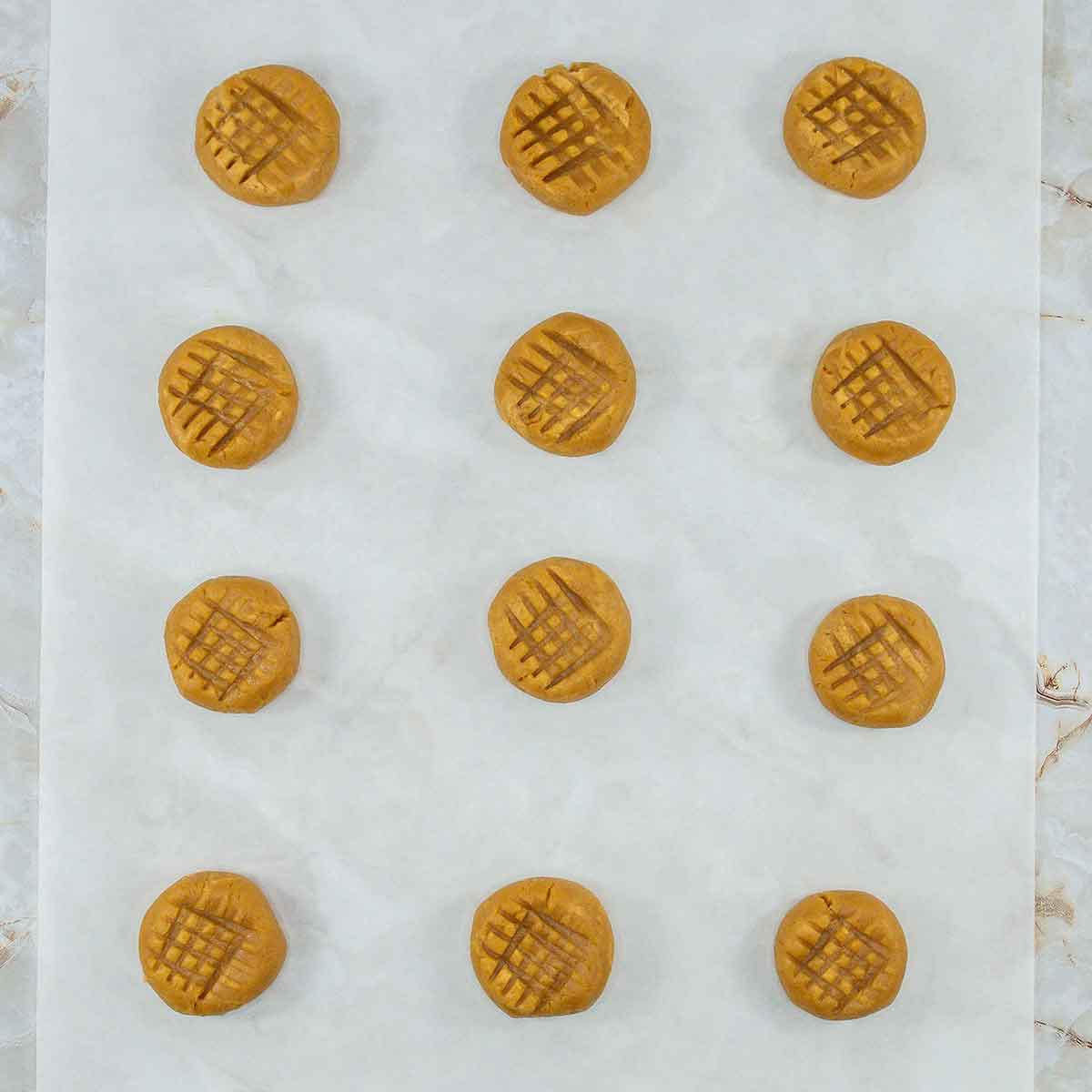 Twelve unbaked 3 Ingredient Peanut Butter Cookies are evenly spaced on a parchment-lined baking sheet, each showcasing a signature crisscross pattern. The scene is set against a light beige marble background, adding an elegant touch to this simple delight.