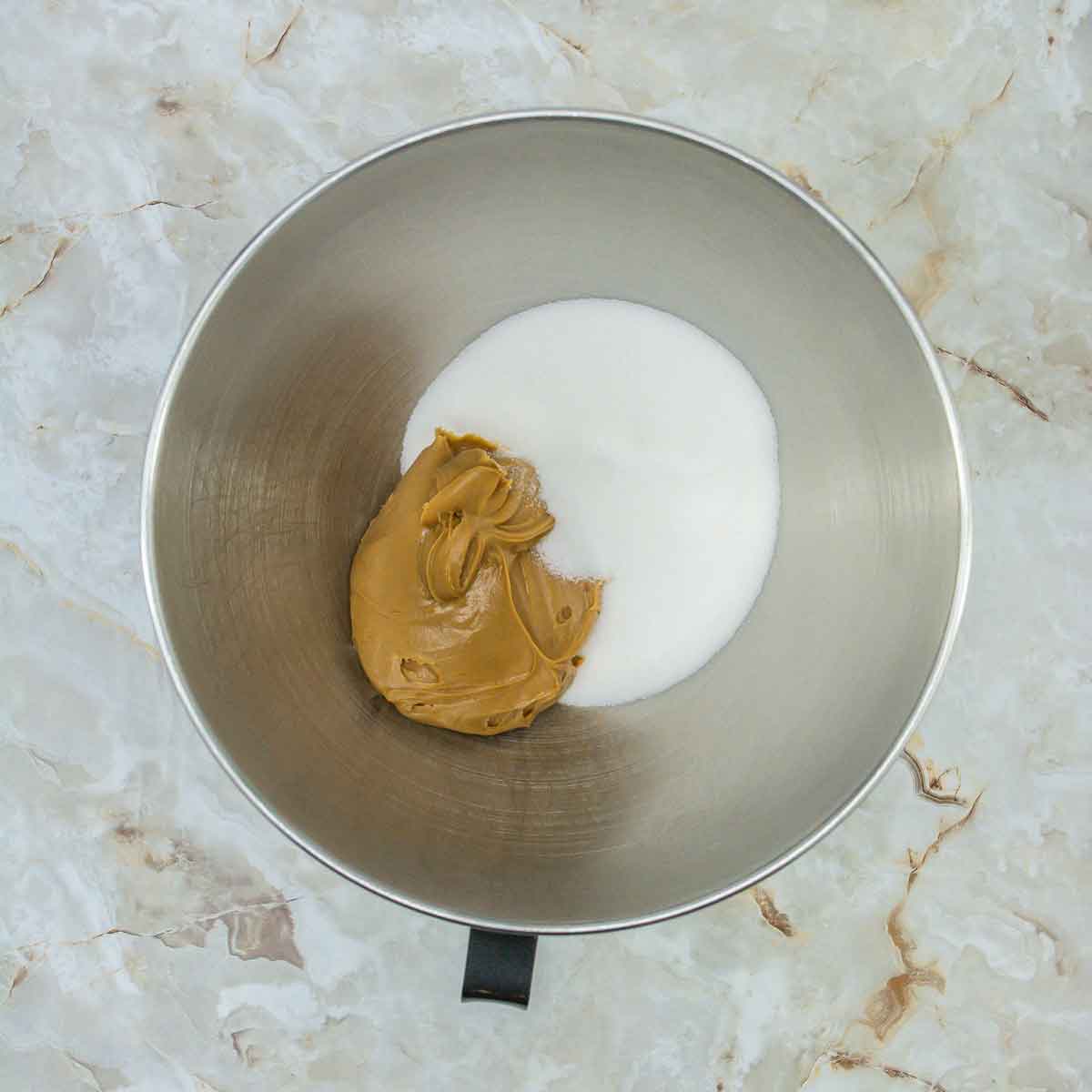 A metal mixing bowl on a marbled surface holds the beginnings of 3 Ingredient Peanut Butter Cookies, with a dollop of peanut butter and a pile of sugar nestled side by side.