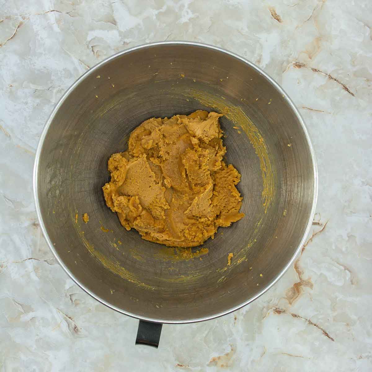 A metal mixing bowl containing a thick, light brown dough for 3-ingredient peanut butter cookies is placed on a marbled countertop.