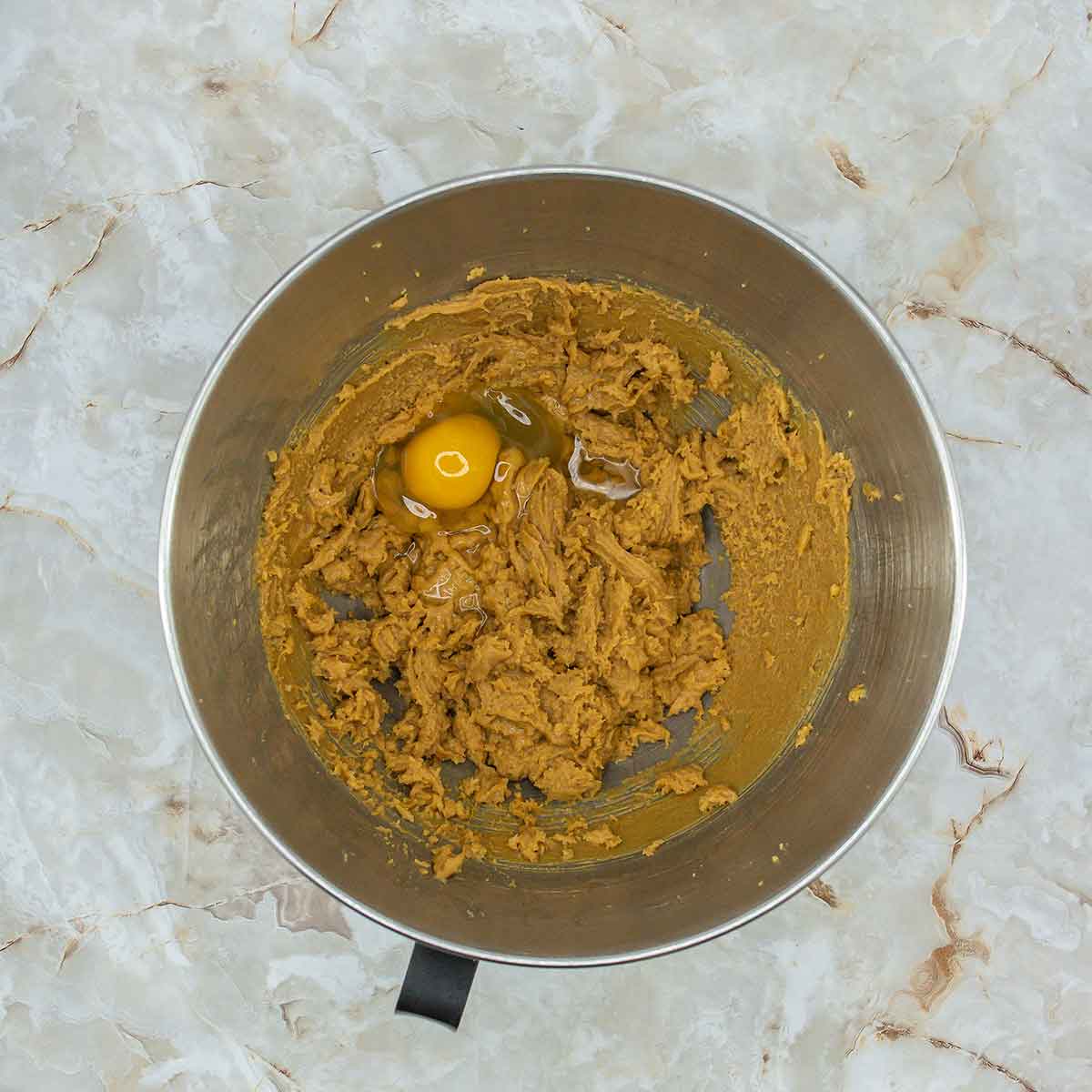 A mixing bowl with cookie dough and a raw egg on top rests on a marbled countertop. The light brown hue suggests a blend of sugar and butter, perfect for crafting 3 Ingredient Peanut Butter Cookies. The intact yolk waits to be blended in, promising delicious simplicity.