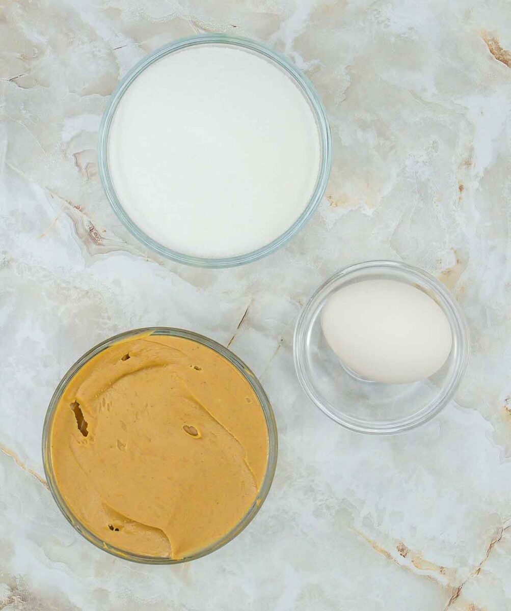 A top view showcases three bowls on a marble surface, perfectly poised to create 3 ingredient peanut butter cookies: one brimming with creamy peanut butter, another filled with granulated sugar, and the last cradling a whole egg.
