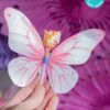 A hand holds a butterfly-shaped card with pink and white wings, wrapped around a colorful lollipop, labeled "template (Copy) (Copy)." The background features more pink decor, and text indicating the design can be edited using Canva.