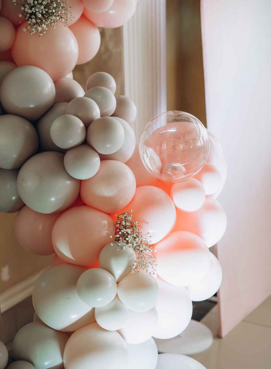 A decorative balloon arrangement featuring clusters of pink, white, and transparent balloons. Small white flowers are interspersed among the balloons, adding a delicate touch to the festive decoration.