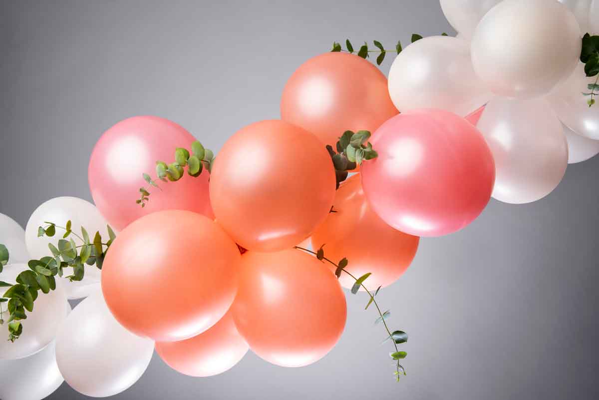 A cluster of orange, pink, and white balloons intertwined with green eucalyptus branches against a gray background. The arrangement appears festive and decorative.