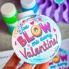 A hand holds a round Valentine tag with "You Blow Me Away Valentine" text, set against a colorful heart-themed backdrop. The background features bubbles and a blurred purple object, with an editable name option included.