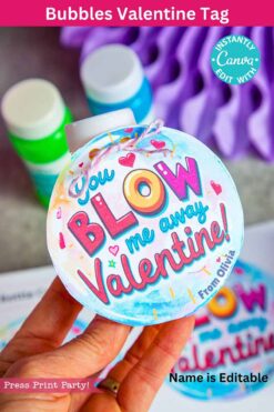 A hand holds a round Valentine tag with "You Blow Me Away Valentine" text, set against a colorful heart-themed backdrop. The background features bubbles and a blurred purple object, with an editable name option included.