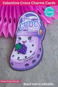A charming Valentine's card showcasing a smiling purple Croc shoe with eyes and a grape charm, alongside the text "You CROC my world!" It includes "Happy Valentine’s Day! From Olivia" at the bottom, set against a pink honeycomb decorative backdrop.