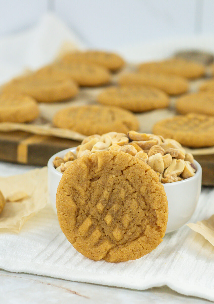 A close-up of a 3 ingredient peanut butter cookie leaning against a small bowl filled with peanuts. In the background, more of these simple cookies are arranged on a wooden tray. The scene is set on a white cloth.