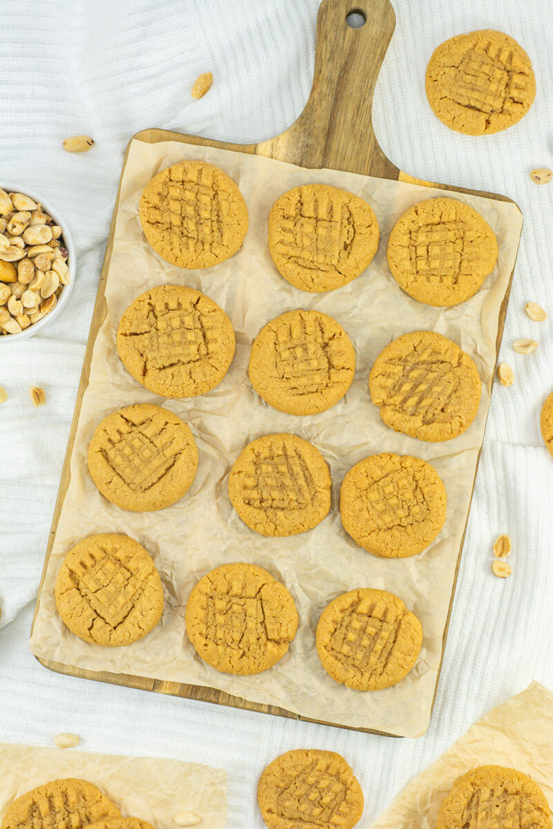 A wooden board holds a dozen delightful 3 Ingredient Peanut Butter Cookies on parchment paper. Neatly arranged, each cookie displays a crisscross fork pattern. Peanuts are scattered around the board, complemented by a small bowl of peanuts to the side.