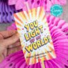 A hand holds the template (Copy) (Copy), a colorful Valentine's card with orange rays and hearts, reading "YOU LIGHT UP MY WORLD! Happy Valentine's Day! From Carson." Attached are two glow sticks, set against pink paper decorations.