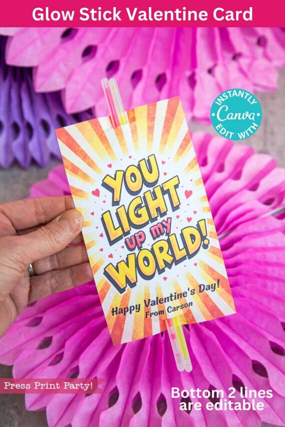A hand holds the template (Copy) (Copy), a colorful Valentine's card with orange rays and hearts, reading "YOU LIGHT UP MY WORLD! Happy Valentine's Day! From Carson." Attached are two glow sticks, set against pink paper decorations.