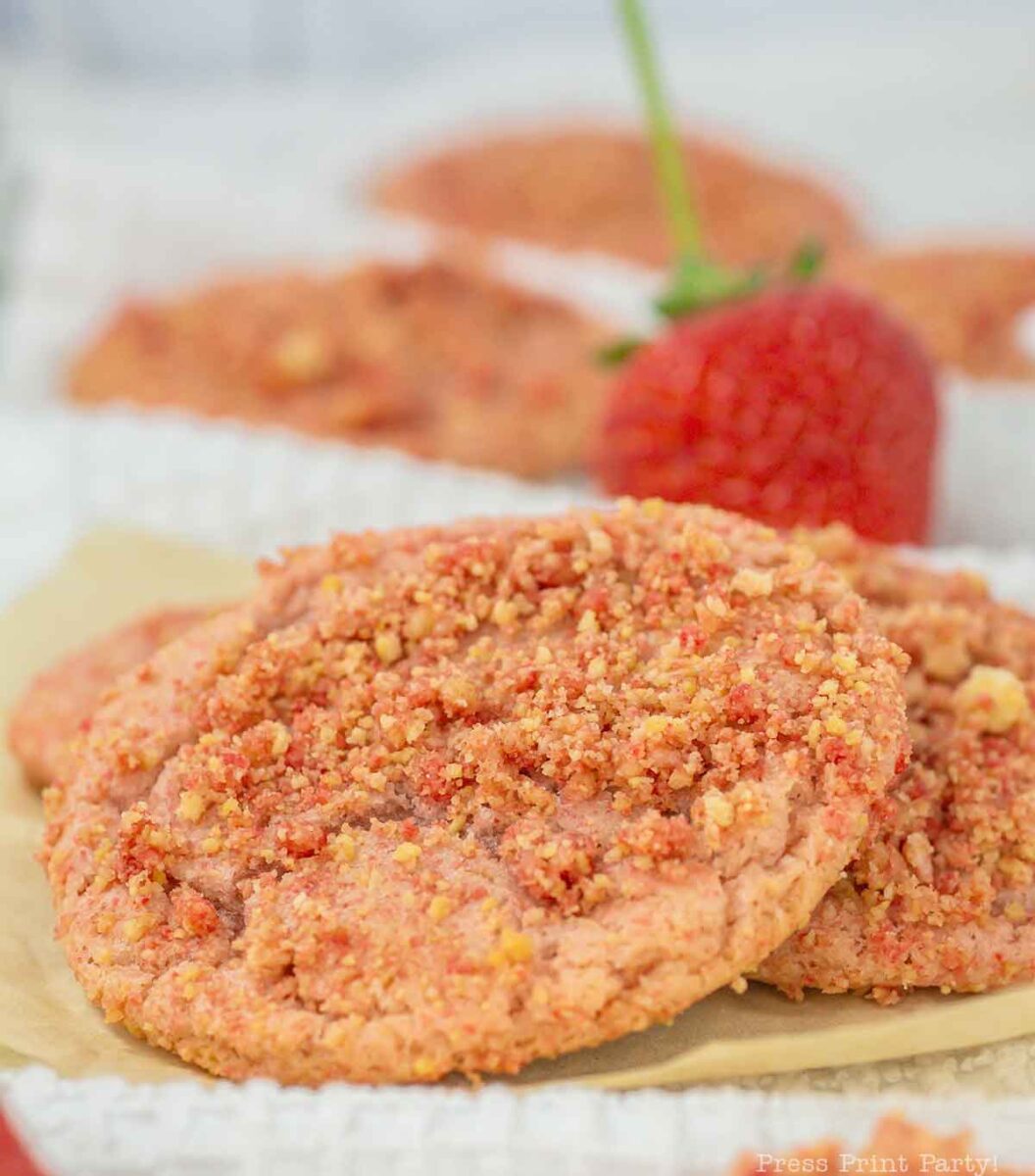 Close-up of strawberry crumble cookies stacked on a sheet of parchment paper, with a blurred fresh strawberry in the background. The cookies are pink with a crumbly texture, suggesting a sweet, fruity flavor.