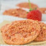 Close-up of strawberry crumble cookies stacked on a sheet of parchment paper, with a blurred fresh strawberry in the background. The cookies are pink with a crumbly texture, suggesting a sweet, fruity flavor.