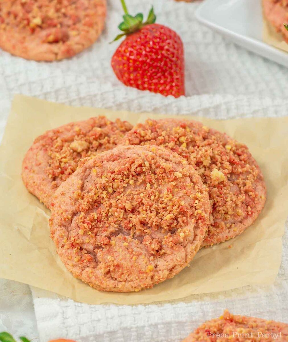 Three strawberry crumble cookies on brown parchment paper. The cookies are topped with a crumbly mixture and are surrounded by whole strawberries on a textured white background.