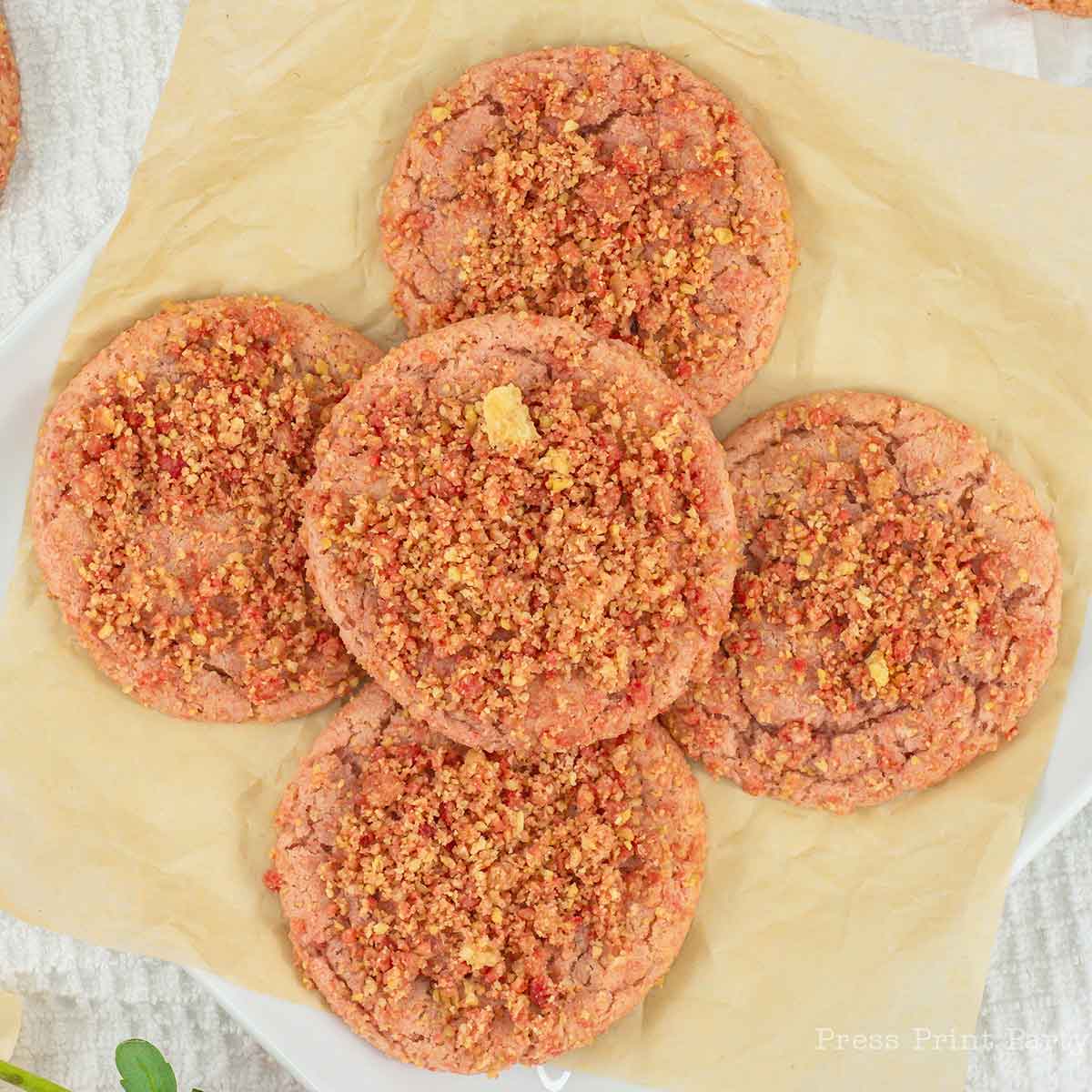 Five strawberry cookies topped with crumbled pieces, arranged in a flower pattern on beige parchment paper. The cookies have a pinkish hue with a crumbly texture.