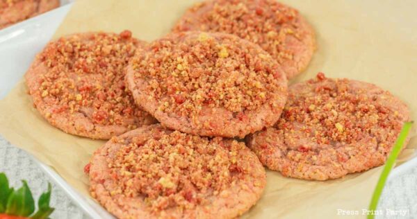 A plate of cookies topped with a crumbly mixture, placed on parchment paper. The cookies have a light pink hue, suggesting a fruity or flavored variation. They are arranged in a slightly overlapping pattern.